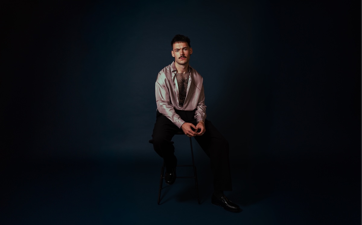 Musician James Marriott sitting on a stool in a satin shirt with a mustache against a dark blue studio background.